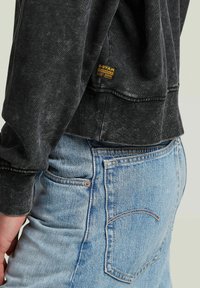 Felpa nera a maniche lunghe con tessuto testurizzato e polsini a coste, con etichetta gialla del logo G-Star; abbinata a jeans azzurri chiari.