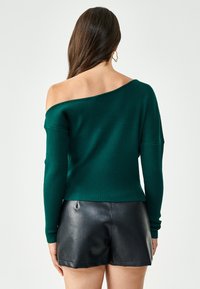 Maglione a coste verde con design off-shoulder, maniche lunghe e vestibilità rilassata, abbinato a pantaloni corti in pelle nera.