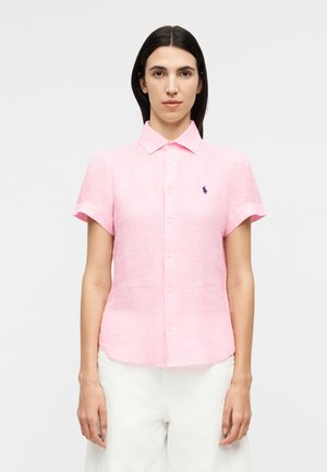 Γυναίκα που φοράει ροζ ανοιχτό ροζ πουκάμισο με κοντό μανίκι, γιακά και κεντημένο λογότυπο, συνδυασμένο με λευκό ψηλόμεσο παντελόνι.