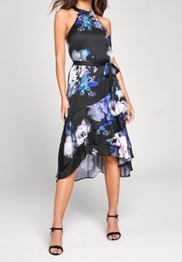Frau trägt ein schwarzes ärmelloses High-Low-Kleid mit blau-weißer Blumenmuster, gebundenem Taillengurt und schwarzen Knöchelriemenabsätzen vor einem grauen Hintergrund.