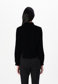 Mustast sametist lühike jakk pikki varrukatega ja kraega, millel on nööbid varrukatel, koos mustade pükstega.