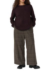 Vrouw in een donkerpaarse lange mouwen trui, brede bruine geruite broek en zwart veterschoenen met handen in de zakken.