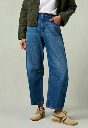 Isik, kes kannab kõrge vöökohaga laia säärega siniseid teksapükse, beežid spordijalatsid, valget särki ja tumerohelist tikandiga jopet, seisab ühe käega taskus.