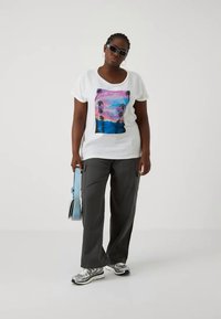 Balta marškinėliai su spalvinga grafika, vaizduojančia palmės ir saulėlydį, derinami su pilkais krosoveriais ir sidabriniais sportiniais bateliais.