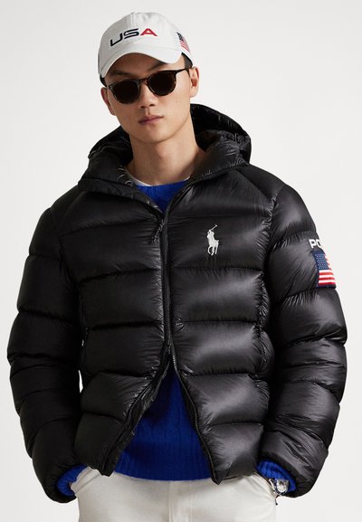 Fekete kapucnis steppelt dzseki bordázott mandzsettákkal, elöl cipzárral és fehér logóval. Kék kötött pulóver fölött viselve.