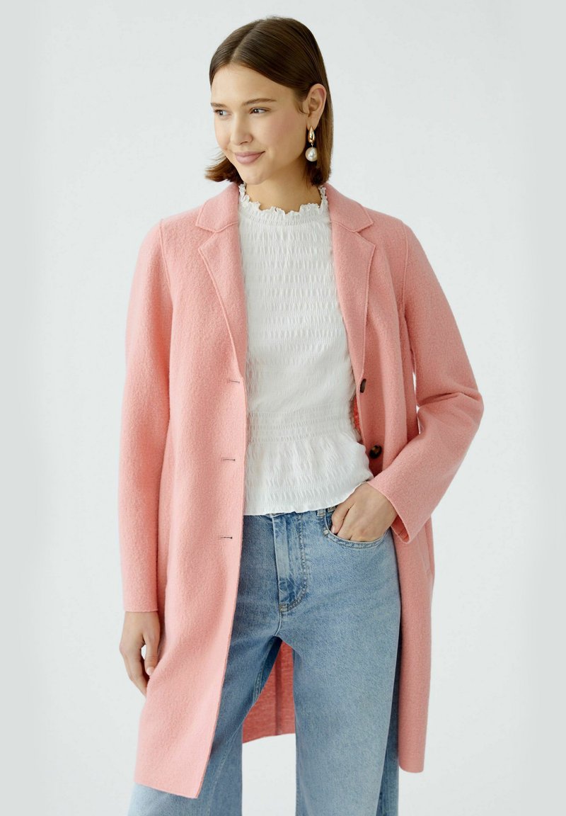 Rosa Wollmantel mit strukturiertem Kragen, drei Knöpfen und taillierter Passform über einem weißen strukturierten Oberteil und hellblauen Denim-Jeans.
