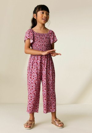 Noor juukseline tüdruk roosakas lillemustrilise kombekaga ja roosakaskuldsetes sandaalides, kätega žeste tegemas, taustaks lihtne beež sein.