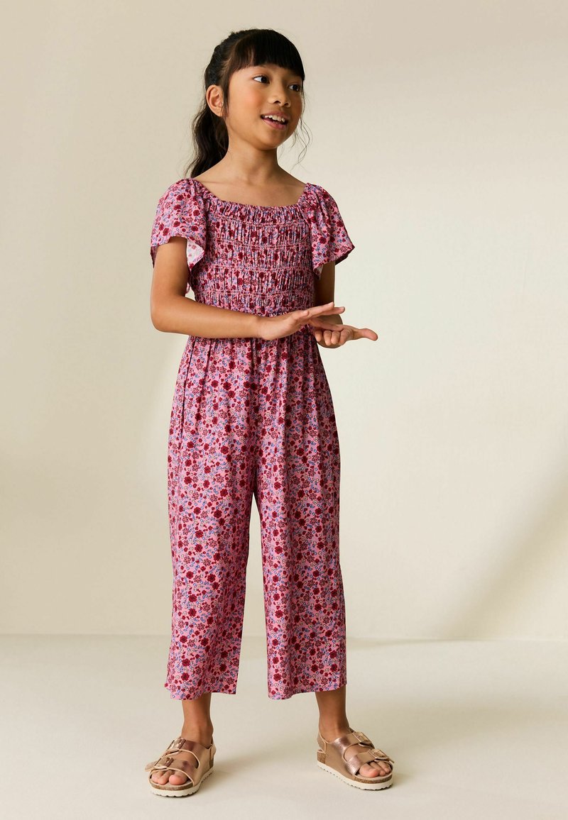 Fiatal lány sötét hajjal, rózsaszín virágmintás overálban és rózsaarany szandálban, kézmozdulatokat téve egyszínű bézs háttér előtt.