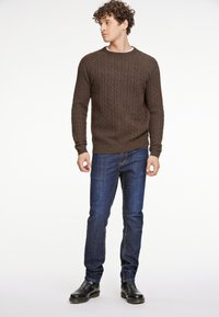 Ruskea kabelikude neule pyöreällä kaula-aukolla, yhdistettynä tummansinisiin farkkuihin ja mustiin nilkkabootseihin. Malli seisoo rentona, kädet sivuilla.