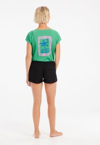 Grön kortärmad skjorta med en färgglad palmträdsgrafik på baksidan, tillsammans med svarta shorts med elastisk midja.