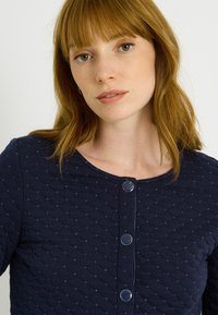 Top acolchado azul marino con patrón de diamantes, con cuello redondo y cuatro botones decorativos en la parte delantera. Textura de tejido suave.