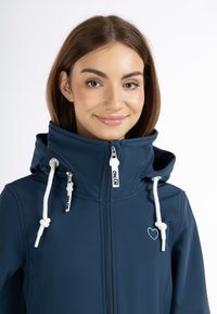 Giacca softshell blu con colletto alto, lacci bianchi, chiusura con zip e dettagli ricamati a forma di cuore. Materiale liscio e resistente all'acqua.