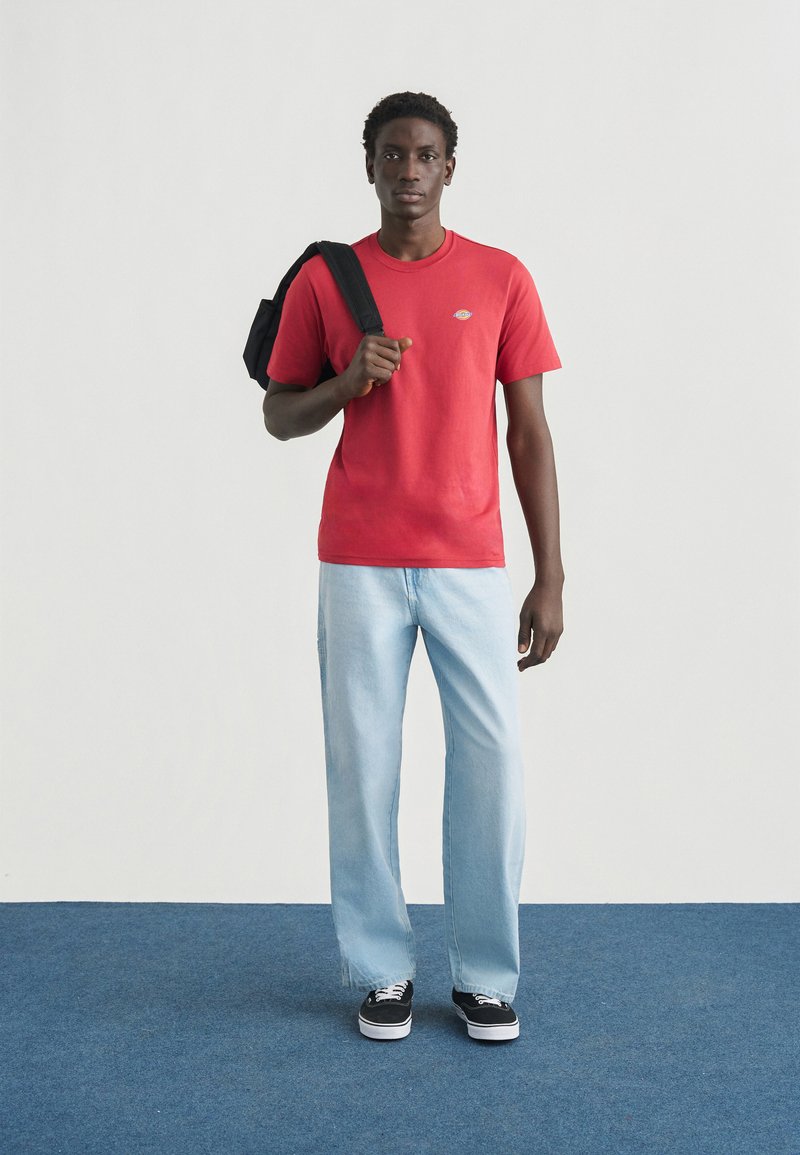 Rood katoenen T-shirt met een klein logo, lichtblauwe rechte jeans en zwarte sneakers. Model draagt een zwarte rugzak over één schouder.