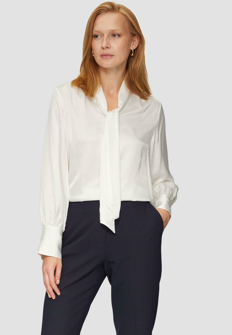 Blouse en soie blanche avec un nœud à l'avant, manches longues avec des poignets froncés, et texture lisse. Associée à un pantalon sombre ajusté.