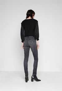 Fekete kötött pulóver áttetsző puff ujjakkal, sötétszürke skinny farmerral és fekete bokacsizmával párosítva, hátulról nézve.