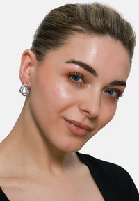 Orecchini a cerchio in argento con design intrecciato, superficie liscia e finitura lucida, indossati da una modella con un top a tonalità neutra e uno sfondo pulito.