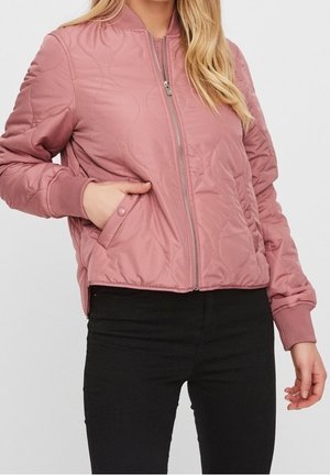 Kobieta ubrana w pikowaną różową kurtkę typu bomber z prążkowanymi mankietami, zamkiem błyskawicznym z przodu i kieszeniami na zatrzaski, w połączeniu z czarnymi spodniami.