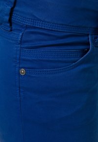Pantaloni in denim blu con una vista ravvicinata dell'area della tasca, caratterizzati da un bottone argento e dettagli di cucitura distintivi sul tessuto.