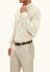 Homme ajustant son col, portant une chemise beige à boutons, un pantalon beige clair et une ceinture marron foncé sur un fond uni.
