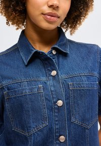 Γυναίκα που φορά μπλε τζιν πουκάμισο με κουμπιά, δύο τσέπες στο στήθος, ορατό γιακά και κοντά σγουρά μαλλιά σε απλό φόντο.