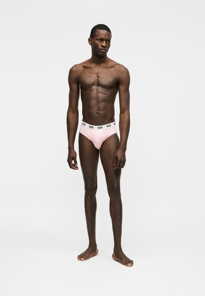 Homme mince à la peau foncée, debout pieds nus devant un fond blanc uni, portant un slip rose pâle avec une ceinture blanche à logo.
