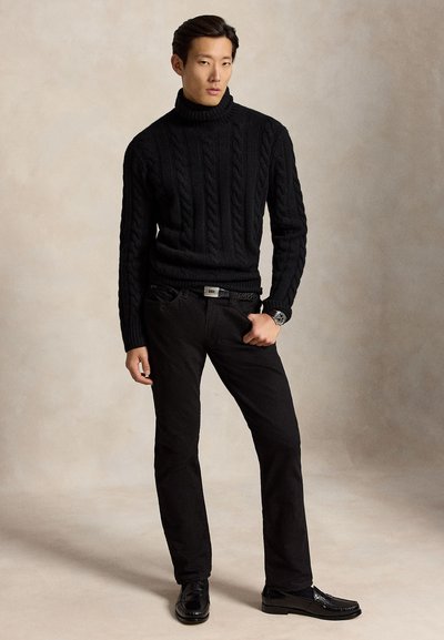 Homme portant un pull à col roulé en maille torsadée noire, un pantalon noir, une ceinture noire, des mocassins noirs et une montre-bracelet, debout devant un fond neutre.