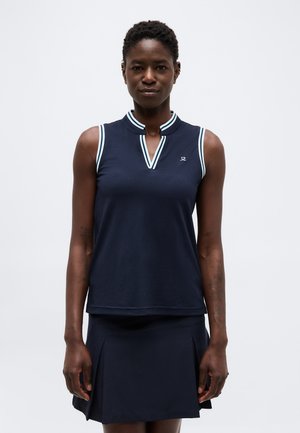 Nő ujjatlan tengerészkék felsőben, fehér szegéllyel és tengerészkék pliszírozott szoknyában, egy egyszínű világosszürke háttér előtt áll.