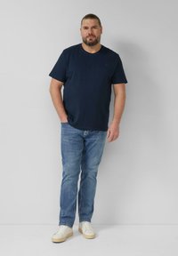 Homme avec une barbe et les cheveux attachés, portant un t-shirt bleu marine, un jean bleu et des baskets blanches, debout devant un fond uni gris clair.