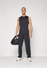Top athletic fără mâneci, de culoare neagră, cu guler rotund, asortat cu pantaloni negri și adidași albi. Modelul ține o geantă de voiaj neagră.