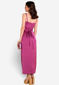 Vestido maxi de satén fucsia con tirantes finos, detalle de cintura atada y una textura suave, combinado con tacones de plataforma dorados y un bolso de lentejuelas.