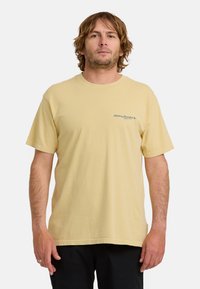 Mężczyzna z średniej długości włosami, noszący jasnoszary t-shirt z krótkim rękawem i czarne spodnie, stojący na tle jednolitego białego tła.