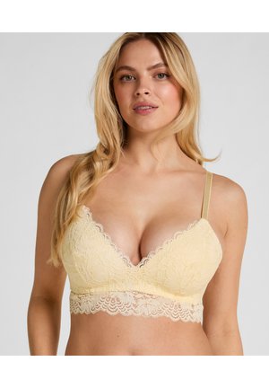 Vrouw die een lichtgele kanten bralette met gegolfde randen en verstelbare dunne bandjes draagt, poseert tegen een effen lichte achtergrond.