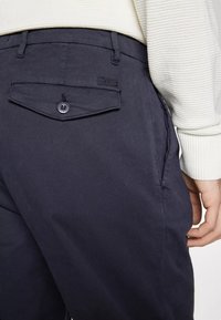 Pantaloni blu navy realizzati in misto cotone, con una tasca posteriore con bottone e patta, e un logo discreto sulla tasca. Texture liscia.
