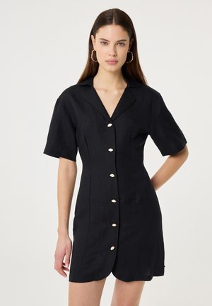 Vrouw met lang bruin haar draagt een zwarte korte mouws jurk met knopen en een getande zoom, en draagt hoepeloorbellen, staand voor een effen achtergrond.