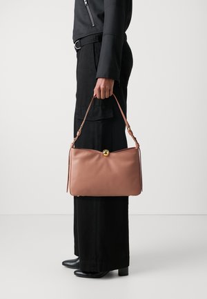 Lys rosa læderhåndtaske med en gylden spænde, rektangulær form, glat tekstur og en aftagelig rem, holdt af en person i sort tøj.