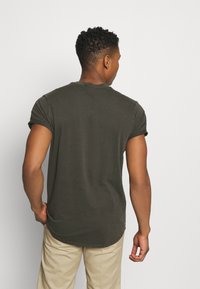 Camiseta ajustada de color verde oliva, con mangas enrolladas y dobladillo redondeado. Material de algodón, textura mínima, sin patrones ni acentos visibles.