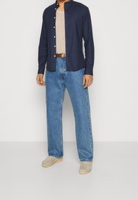 Camicia blu scuro con colletto alla mandarina, maglietta lavorata color crema, jeans azzurri a taglio morbido e scarpe beige senza lacci.