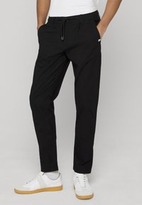 Homme portant un pantalon noir à cordon et des baskets blanches avec semelles en caoutchouc, debout les mains dans les poches, sur un fond uni.