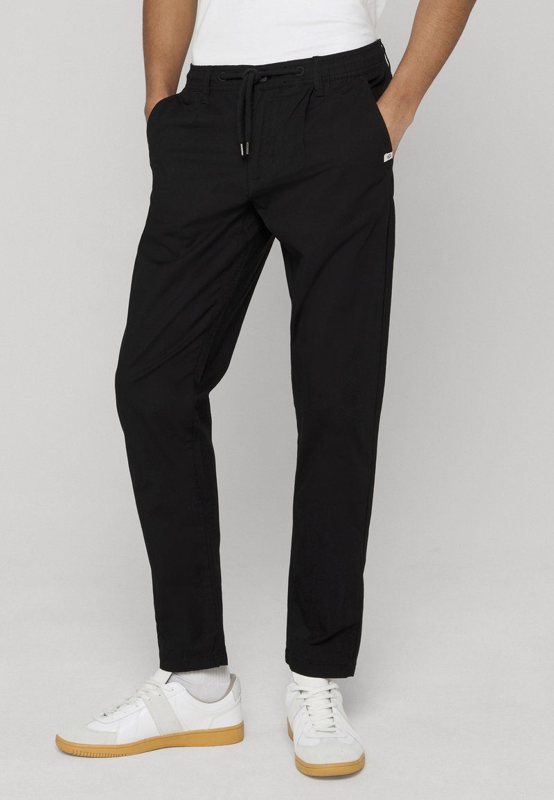 Homme portant un pantalon noir à cordon et des baskets blanches avec semelles en caoutchouc, debout les mains dans les poches, sur un fond uni.