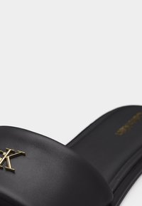 Μαύρο σανδάλι με φάσα που φέρει το χρυσό λογότυπο CK και μαλακό πέλμα, εμφανίζεται σε λευκό φόντο.