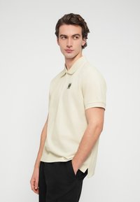 Φωτεινό μπεζ πόλο μπλουζάκι από υφασμάτινη υφή, με κλασικό γιακά και κοντά μανίκια, με διακριτικό λογότυπο στο στήθος.