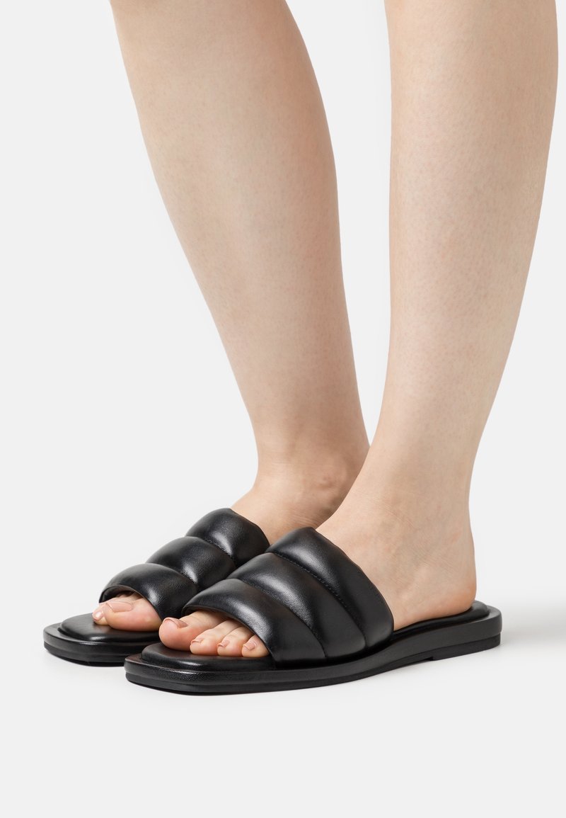 Sandales à glisser en cuir noir avec un design supérieur rembourré, semelle plate et bout ouvert, mettant en valeur une texture lisse et un style minimaliste.