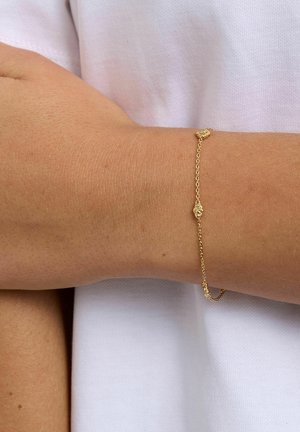 Sart guldkædearmbånd med små teksturerede vedhæng båret på et håndled med lys brun hud, parret med en hvid stofbaggrund.