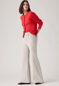 Cardigan de malha vermelha com decote redondo e fecho de botões, combinado com jeans beje de pernas largas e sapatos pretos de bico fino.