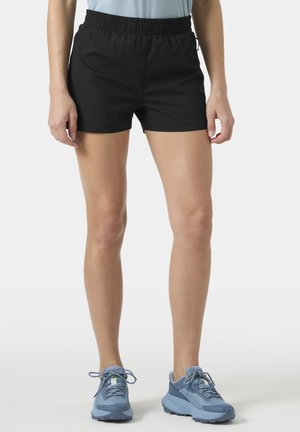 Persoon draagt zwarte shorts met elastische taille en lichtblauwe sportschoenen, staand tegen een witte achtergrond.