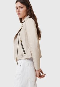 Giacca da motociclista in pelle beige con cerniere nere, design aderente e hardware minimale. Presenta una leggera lucentezza e una texture morbida.