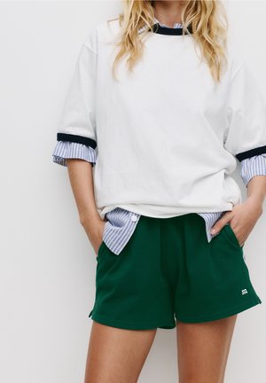 Person iført en hvid oversized skjorte med stribede manchetter lagdelt over en stribet skjorte med knapper og mørkegrønne shorts med hænderne i lommerne.