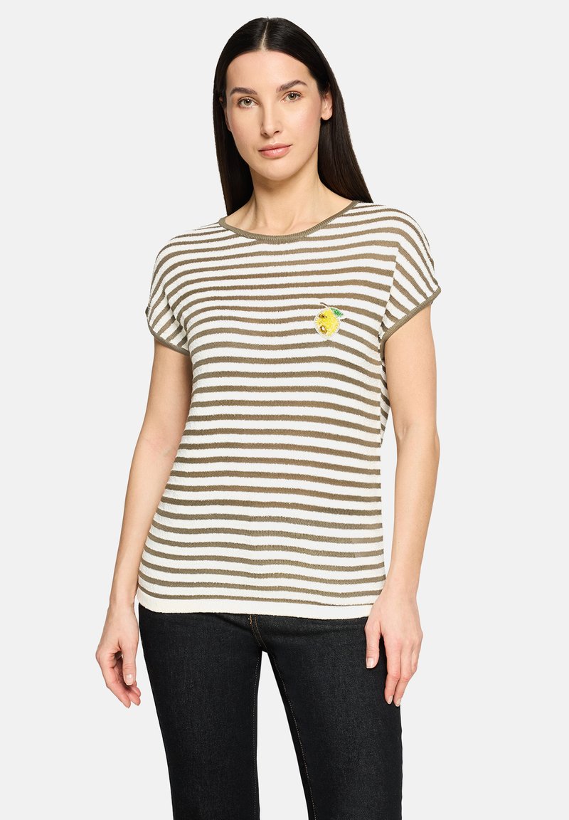 Kvinde med glat mørkt hår iført en kortærmet hvid og olivengrønstribet top med en lille citronpatch på brystet og sorte bukser.