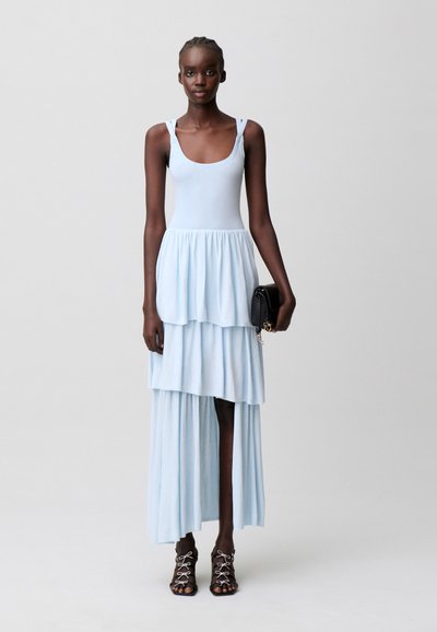 Donna alta che indossa un vestito smanicato azzurro chiaro a balze, tiene una pochette nera e indossa sandali neri con lacci.