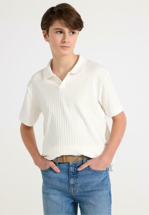 Teini-ikäinen poika, jolla on lyhyet ruskeat hiukset, valkoinen ribbineuloksinen lyhythihainen poolopaita ja siniset farkut, kädet taskuissa.